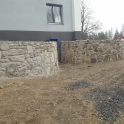 Dwa półokrągłe mury z kamienia polnego, tworzące wejście do budynku z oknem zabezpieczonym folią, widoczne ślady prac ziemnych i gruz.