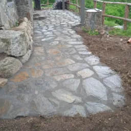 Ścieżka z nieregularnych kamieni ułożona przy kamiennym budynku, z widocznymi elementami drewnianej balustrady i świeżo obsypaną ziemią po bokach.