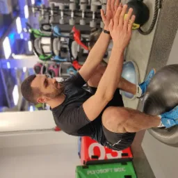 Mężczyzna z brodą ćwiczy mięśnie brzucha na piłce gimnastycznej w siłowni, w tle widoczne obciążenia i skrzynia plyometryczna.