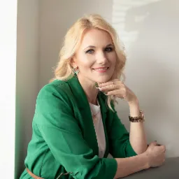 Portret uśmiechniętej blondynki w zielonym żakiecie, opierającej brodę na dłoni, z widocznym złotym zegarkiem i kolczykami w kształcie gwiazd; w tle jasna ściana z delikatnymi cieniami.