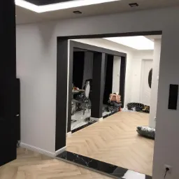 Nowoczesne wnętrze z jasną podłogą ułożoną w jodełkę, czarne ramy drzwiowe i listwy przypodłogowe z czarnego marmuru, oświetlenie LED na suficie.