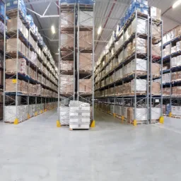 Panorama wnętrza magazynu wysokiego składowania z rzędami metalowych regałów wypełnionych kartonami, zabezpieczonymi folią stretch, oraz paletami z towarem.