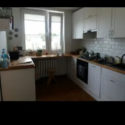 Jasna kuchnia z białymi szafkami, drewnianym blatem i białymi płytkami metra na ścianie, widoczne okno z zasłonami i drewniany stołek.