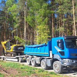 FELEK - Niebieska ciężarówka Renault z żółtą koparką JCB na lawecie, na tle leśnej drogi. Dzień słoneczny, widoczne drzewa i fragment nieba. Rejestracja OKL 57128.