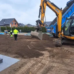 Żółto-niebieska koparka JCB 8080 i pracownicy wyrównują teren pod budowę domu jednorodzinnego. Widoczne ślady opon i świeżo rozplantowana ziemia.