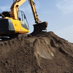 FELEK - Żółto-czarna koparka JCB z uniesionym ramieniem i łyżką, wysypująca ziemię na dużą pryzmę. W tle błękitne niebo i zieleń. Praca w toku.