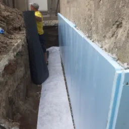 Ocieplanie fundamentów: pracownik układa membranę kubełkową przy ścianie fundamentowej zabezpieczonej styropianem, widoczny wykop i biała geowłóknina.