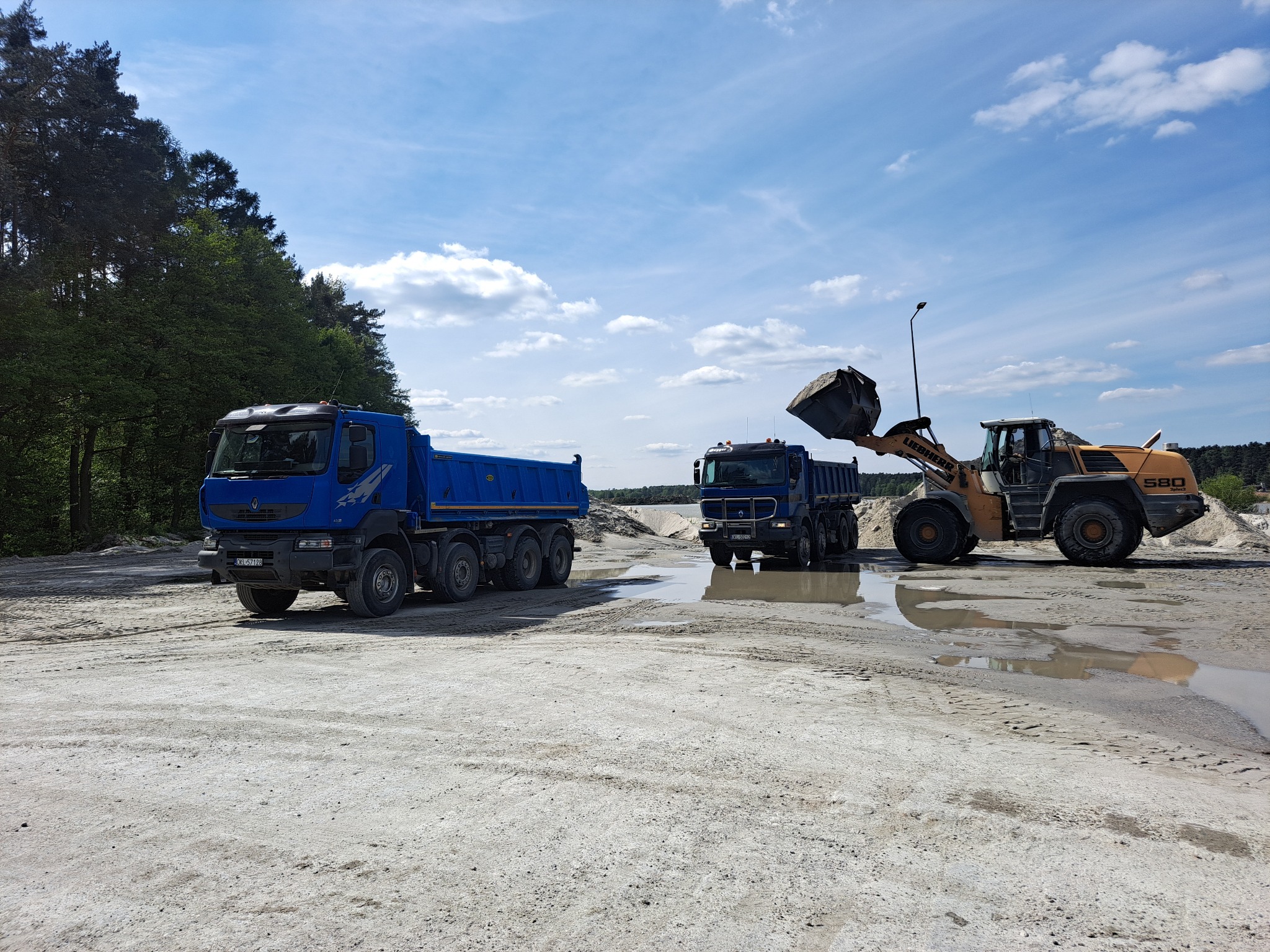 Żółta ładowarka Liebherr 580 nabiera urobek do niebieskiej wywrotki Renault na placu budowy. W tle druga wywrotka i zalesiony teren pod błękitnym niebem.
