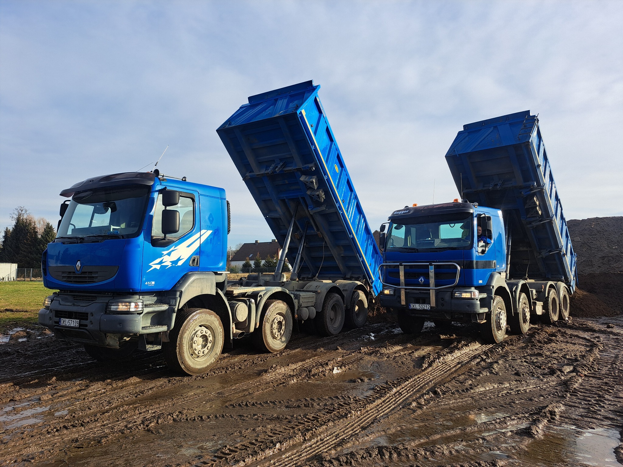 Dwa niebieskie wywrotki z podniesionymi skrzyniami na błotnistym terenie. Widoczne ślady opon i sterta ziemi w tle. Słoneczny dzień, błękitne niebo z chmurami.