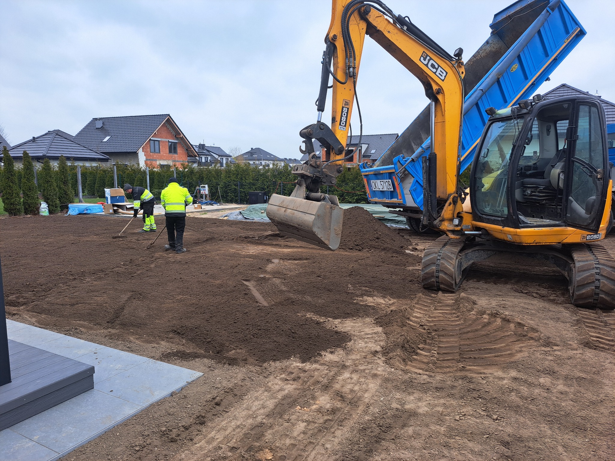 Żółto-niebieska koparka JCB 8080 i pracownicy wyrównują teren pod budowę domu jednorodzinnego. Widoczne ślady opon i świeżo rozplantowana ziemia.