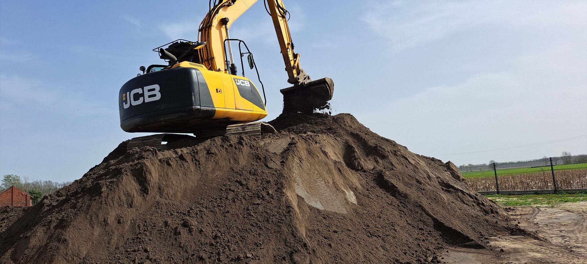 Żółto-czarna koparka JCB z uniesionym ramieniem i łyżką, wysypująca ziemię na dużą pryzmę. W tle błękitne niebo i zieleń. Praca w toku.