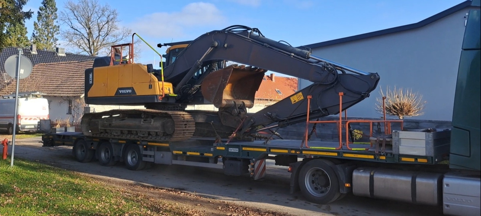 Żółto-czarna koparka Volvo na niskopodwoziowej platformie transportowej, widoczna z boku na tle zabudowań i błękitnego nieba. Transport specjalistyczny.