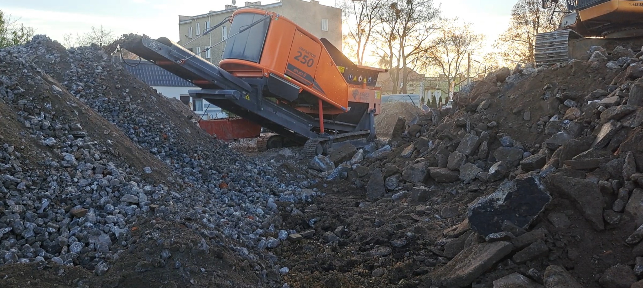 Pomarańczowa kruszarka IMPACTOR 250 EVO II na gąsienicach, otoczona gruzem i ziemią. W tle budynki i drzewa w słoneczny dzień. Widok z dołu.