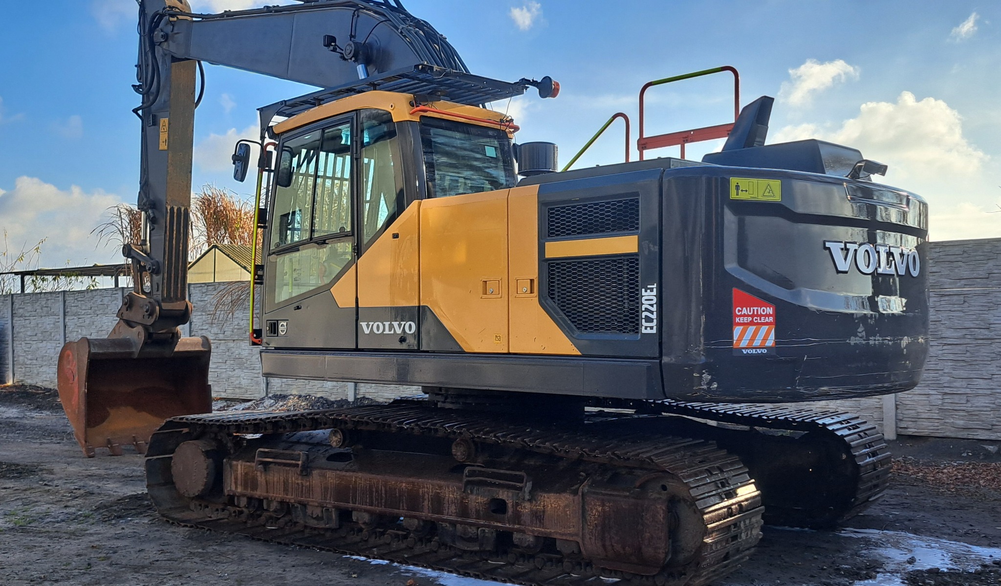 Żółto-czarna koparka Volvo EC220EL na gąsienicach, z łyżką skierowaną w lewo, na tle błękitnego nieba i betonowego ogrodzenia. Widoczne detale: kabina, gąsienice, logo Volvo.