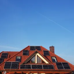 Czerwony dach domu z zamontowanymi panelami słonecznymi w układzie pionowym i poziomym, widoczne okna dachowe i komin z cegły, na tle błękitne niebo z śladami kondensacyjnymi samolotu.