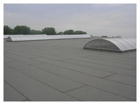 Płaski dach pokryty papą termozgrzewalną w szarym kolorze, widoczne świetliki kopułowe, pochmurne niebo w tle.