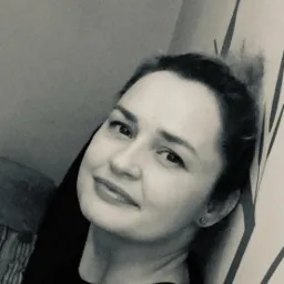 Portret kobiety w czarno-białej tonacji, opierającej się o ścianę z abstrakcyjnym wzorem, uśmiechająca się delikatnie do kamery.