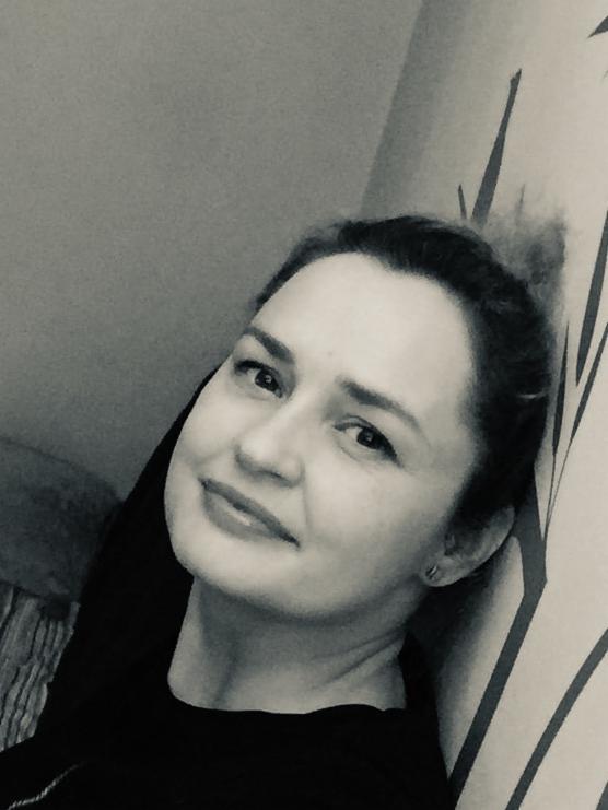 Portret kobiety w czarno-białej tonacji, opierającej się o ścianę z abstrakcyjnym wzorem, uśmiechająca się delikatnie do kamery.