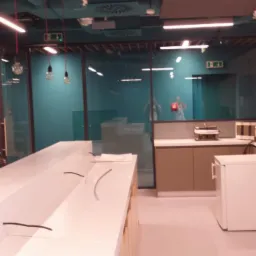 Nowoczesna kuchnia biurowa z długim, białym blatem, szklanymi ścianami działowymi i turkusowymi elementami dekoracyjnymi.