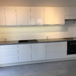 Minimalistyczna kuchnia z białymi, lakierowanymi frontami, szarym blatem i podświetleniem LED pod szafkami wiszącymi, z płytą indukcyjną i piekarnikiem w zabudowie.