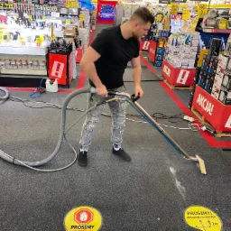 Mężczyzna w czarnej koszulce i jeansach czyści szary dywan w sklepie za pomocą profesjonalnego odkurzacza piorącego; widoczne oznaczenia 'Prosimy! Zachowaj odstęp 2m' na podłodze.