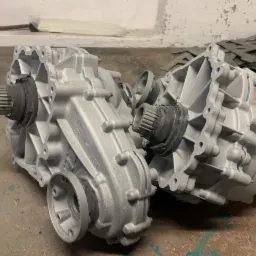 Dwa odświeżone, srebrne korpusy przekładni leżące na szarej powierzchni, z widocznymi detalami konstrukcyjnymi i elementami gumowymi. W tle ściana z odpadającym tynkiem i stos wyciętych elementów...