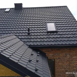 Nowy, czarny dach z blachy trapezowej z oknem dachowym i kominem wentylacyjnym, widok z dołu na fragment budynku z cegły.
