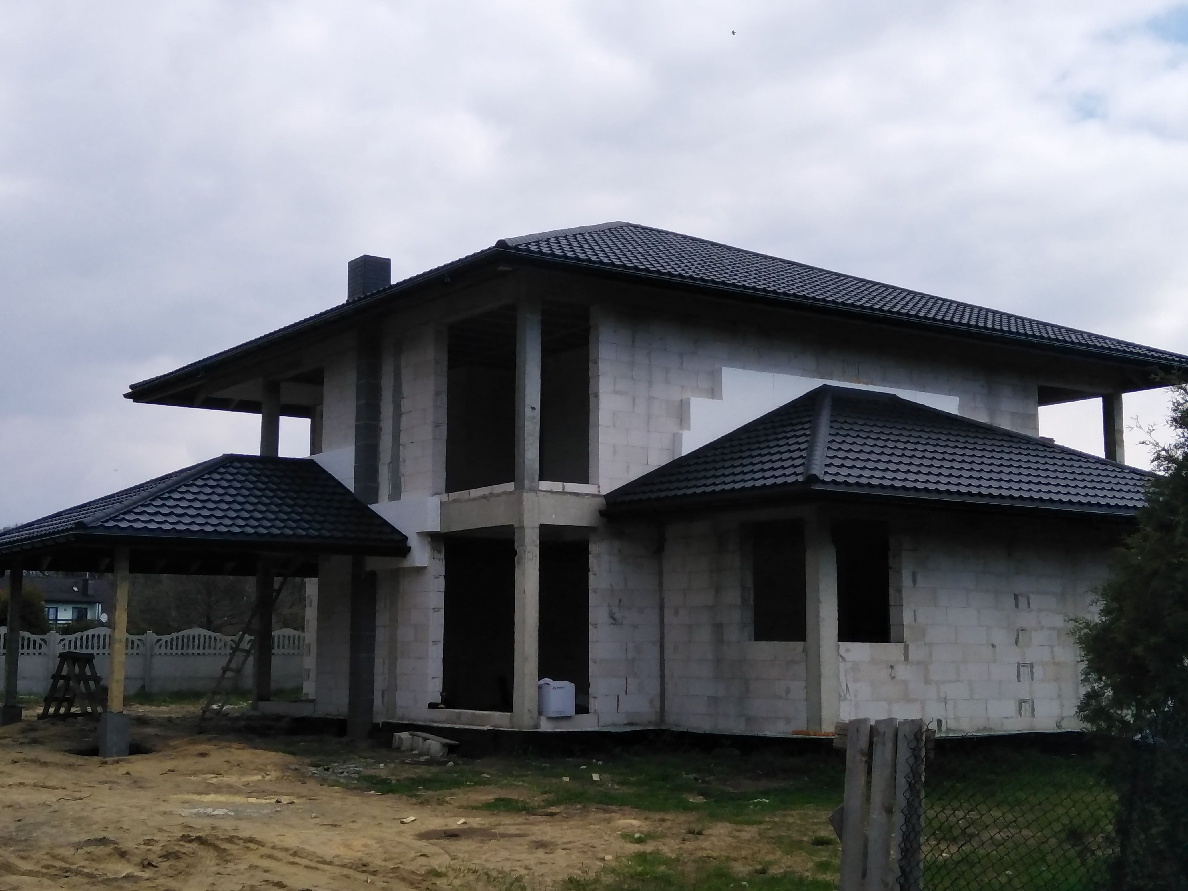 Niedokończony dom z czarnym dachem z blachodachówki, widoczne pustaki i brak okien, zadaszenie nad tarasem również z czarną blachodachówką.