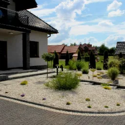 Nowoczesny ogród przydomowy w Krakowie z białym żwirem, niskimi nasadzeniami i strzyżonymi drzewkami iglastymi, widok z poziomu podjazdu z kostki brukowej.