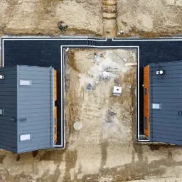 Widok z góry na dwa modułowe domy w trakcie budowy, otoczone wykopami i przygotowanym podłożem, z fragmentem ułożonej kostki brukowej między nimi.