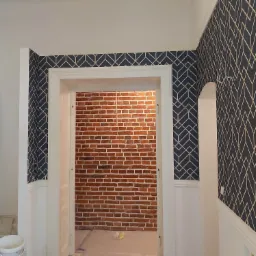 Wnętrze w trakcie remontu: ściany z tapetą w geometryczne wzory, widoczna ściana z czerwonej cegły przez otwarte drzwi, farby i narzędzia malarskie w rogu.