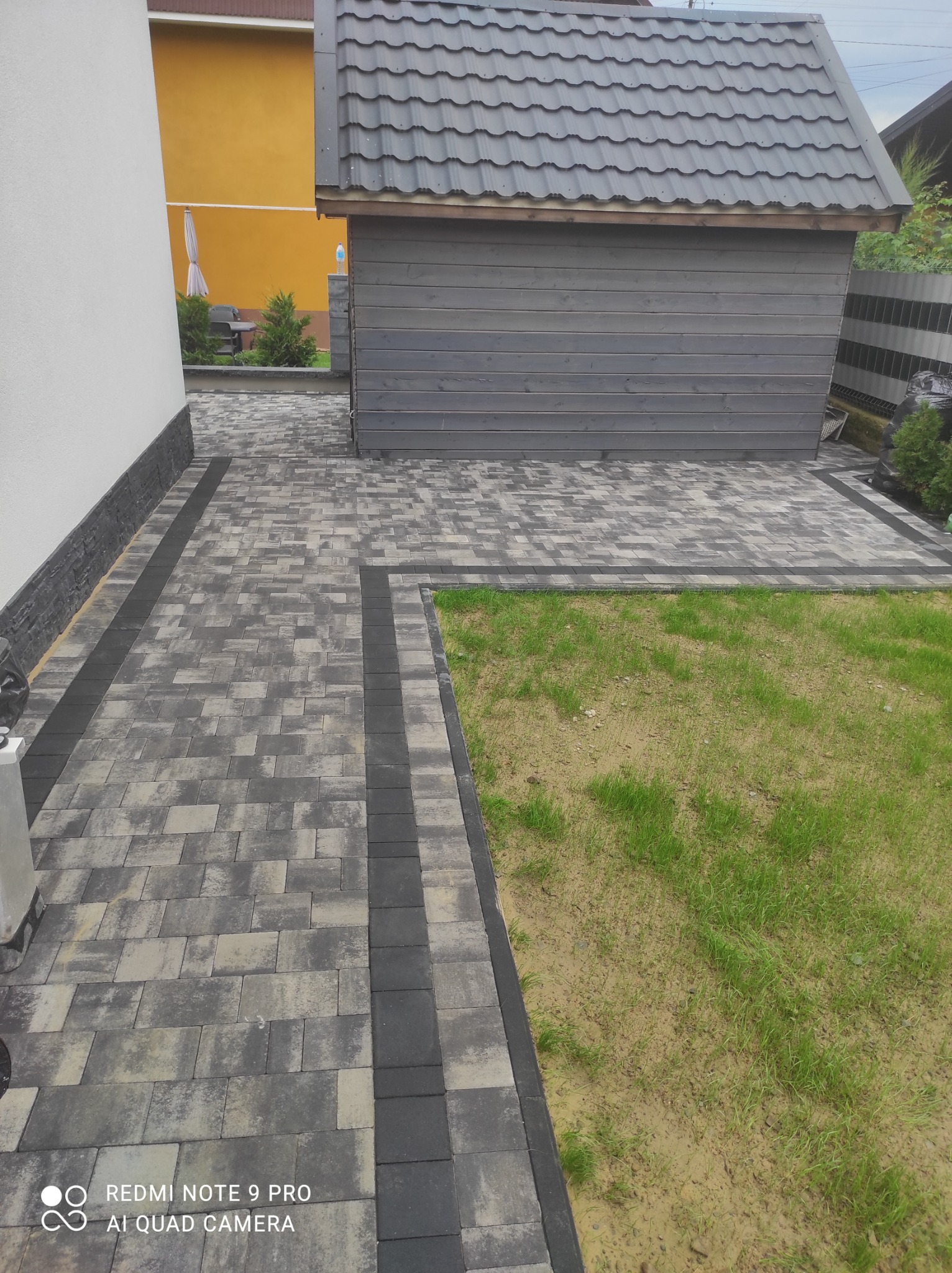 Ułożona ścieżka z szarej kostki brukowej z ciemnoszarym obramowaniem, prowadząca do szarego, drewnianego domku narzędziowego; trawnik z rzadką trawą po prawej stronie.