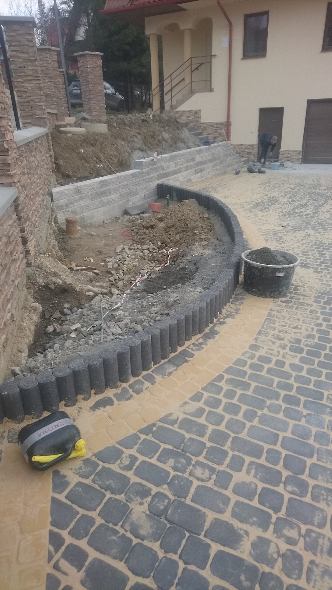 Układanie kostki brukowej w dwóch kolorach, obrzeże z palisady betonowej, przygotowanie terenu pod rabatę, widoczny kanister z płynem do czyszczenia kostki.