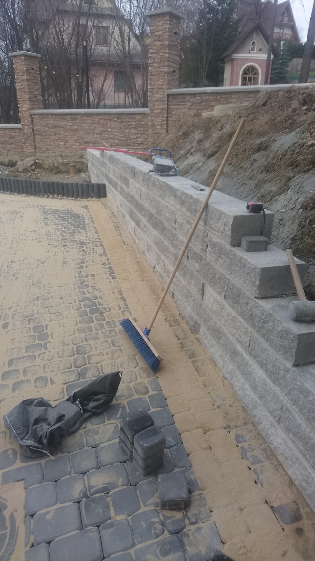 Układanie kostki brukowej z szarej kostki na podsypce piaskowej, z widocznym fragmentem muru oporowego z szarego kamienia i narzędziami: miotła, gumowy młotek, poziomica laserowa.