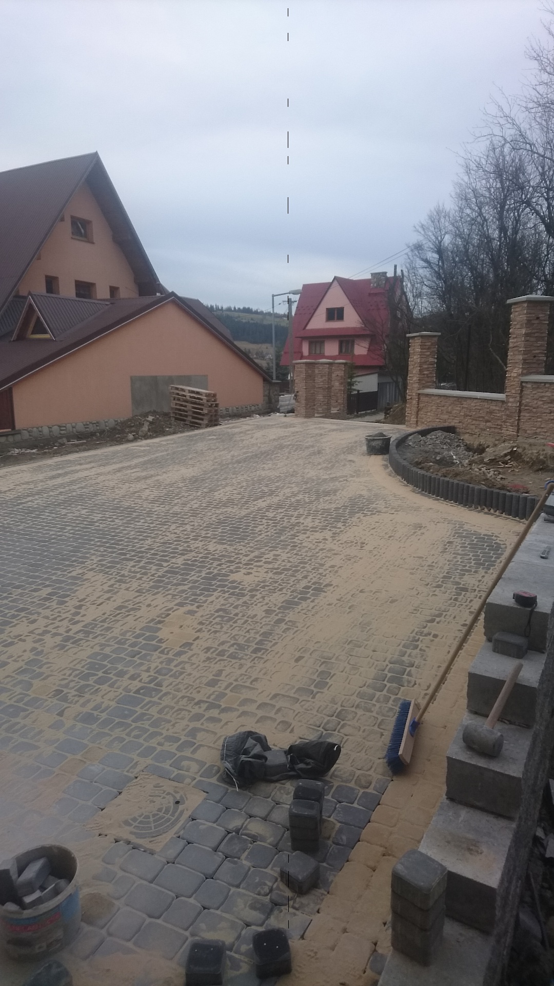 Ułożona kostka brukowa na podjeździe domu, zasypana piaskiem, z widocznymi narzędziami pracy: szczotką i młotkiem brukarskim.