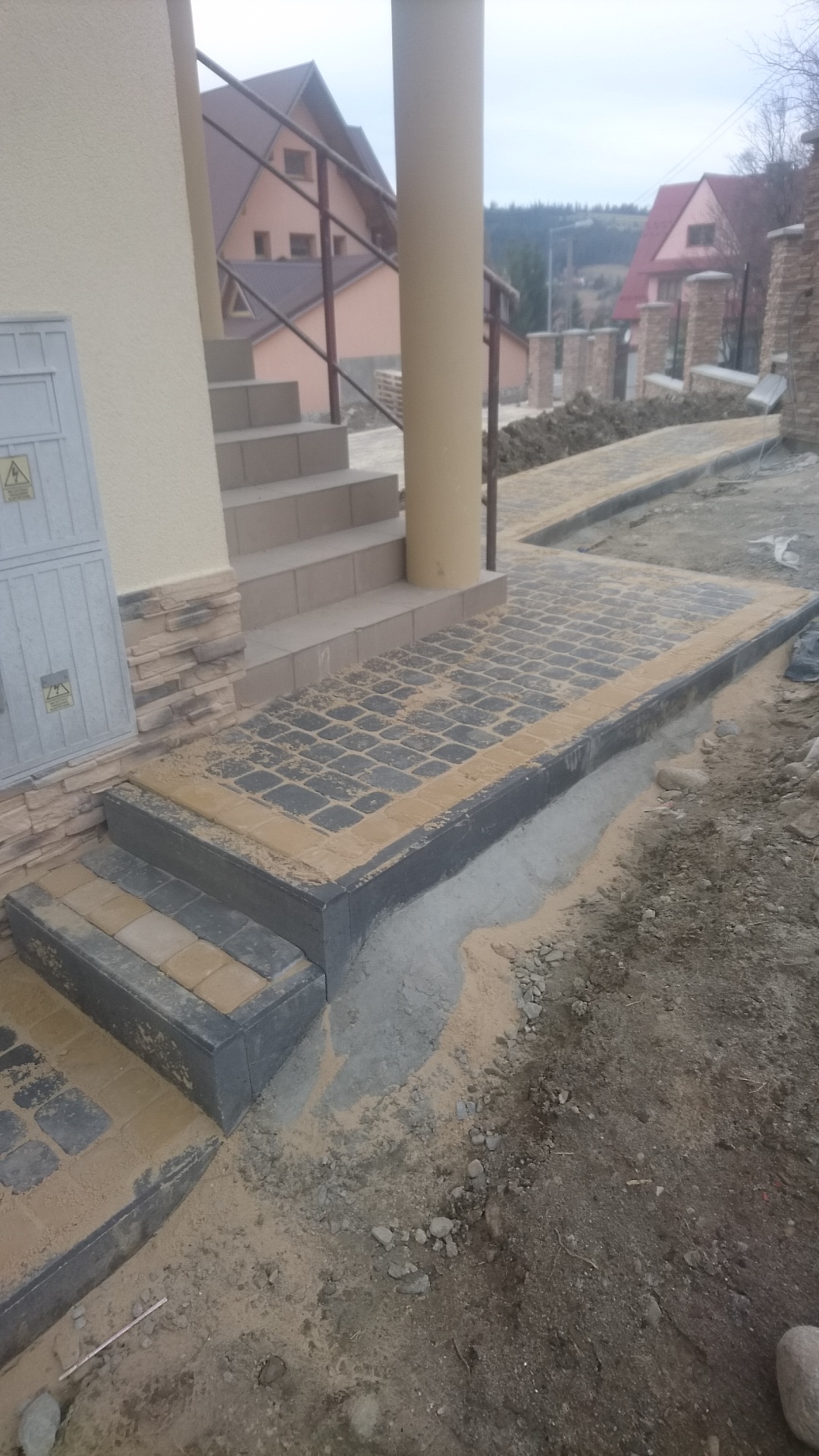 Schody wejściowe i fragment chodnika z kostki brukowej w trakcie prac wykończeniowych, piasek na powierzchni, widoczne budynki w tle.