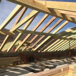 Konstrukcja drewniana więźby dachowej w trakcie montażu, widoczne elementy muru i krajobraz górski w tle.