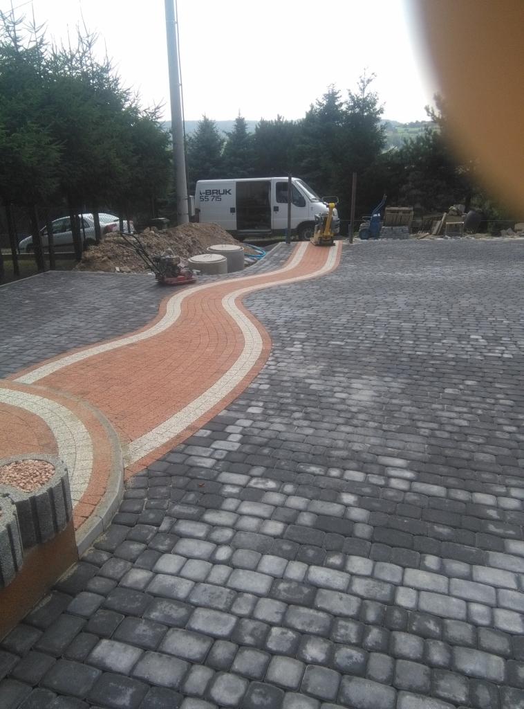 Ułożona nawierzchnia z kostki brukowej w kolorach szarości i czerwieni, z widocznymi maszynami do zagęszczania gruntu i samochodem dostawczym w tle.