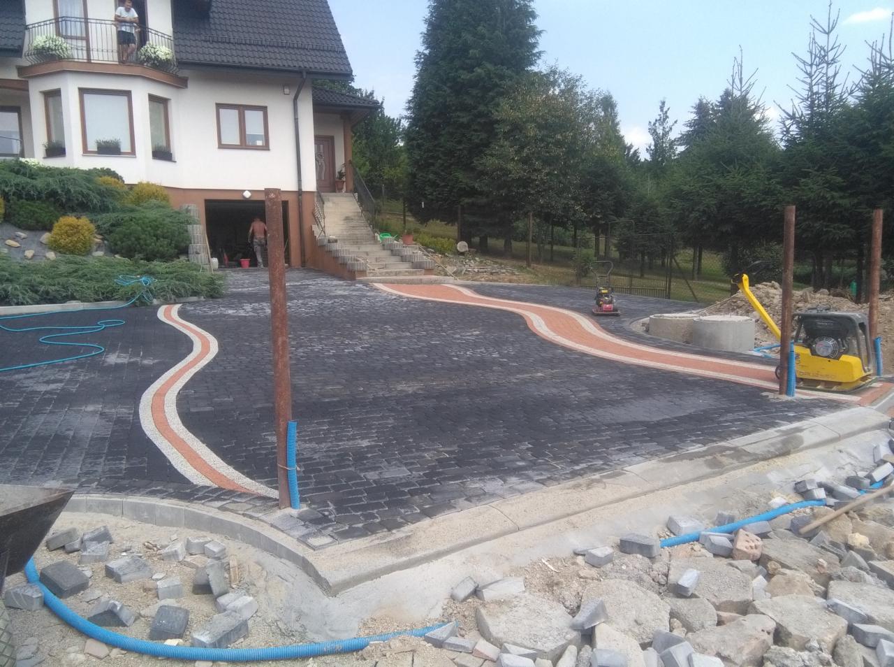 Nowa, czarno-pomarańczowa nawierzchnia z kostki brukowej przed domem jednorodzinnym, widoczne maszyny do ubijania, krawężniki w trakcie montażu, częściowo ułożona, z elementami wykończeniowymi w toku.