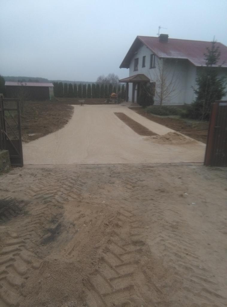Przygotowana podbudowa z piasku pod przyszły podjazd do domu jednorodzinnego, widoczne ślady opon ciężkiego sprzętu, w tle dom z czerwonym dachem i mieszalnik do betonu.