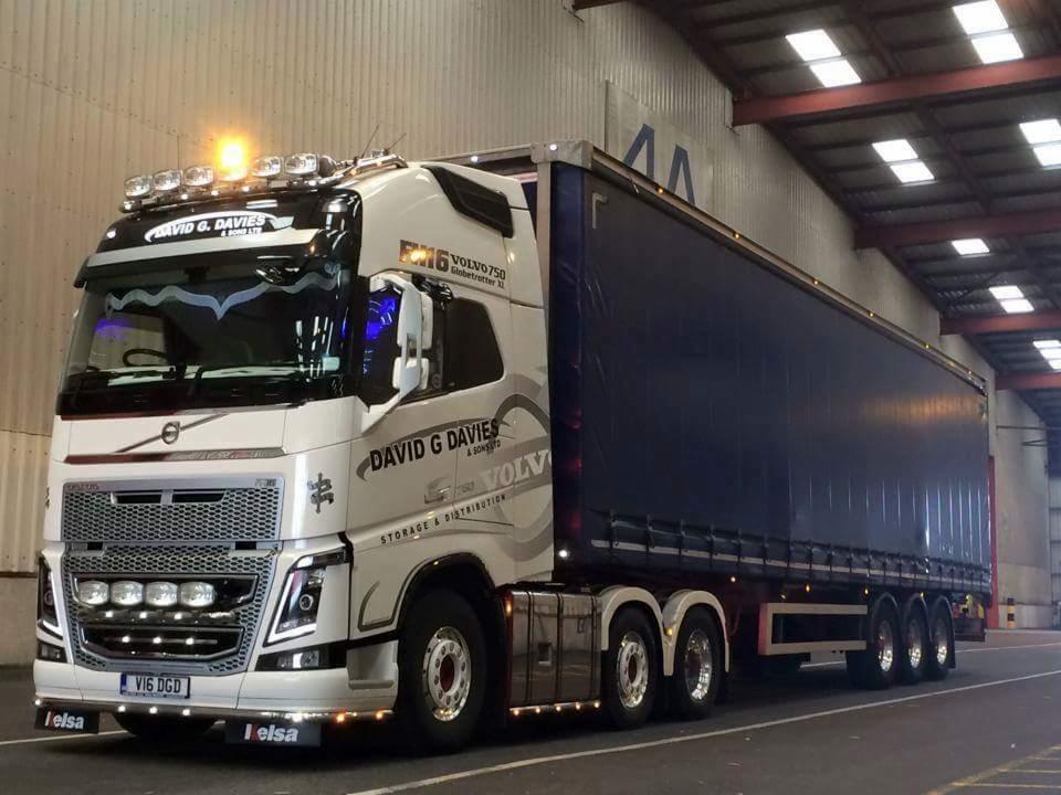 Ciągnik siodłowy Volvo FH z naczepą kurtynową firmy David G Davies Storage & Distribution zaparkowany w hali magazynowej, oświetlenie robocze włączone.