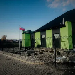 Budynek pomalowany na szaro i jasnozielono z nowym ogrodzeniem panelowym w kolorze czarnym i flagą Polski na maszcie.