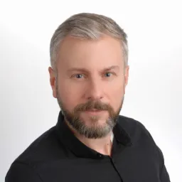 Portret mężczyzny z siwiejącą brodą i włosami, ubranego w czarną koszulę na białym tle.