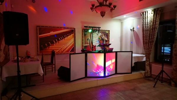 Sala bankietowa z przygotowanym stanowiskiem DJ-a: biały parawan z kolorowym podświetleniem, głośnik na statywie, dekoracje w postaci obrazów i nastrojowego oświetlenia.