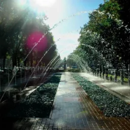 Aleja z fontannami tryskającymi łukami wody, wyłożona cegłą i żwirem, otoczona drzewami w słoneczny dzień.