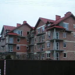 Wielopiętrowy budynek mieszkalny w trakcie budowy, z widoczną surową cegłą i częściowo zamontowanymi oknami, sfotografowany zza ogrodzenia w pochmurny dzień.