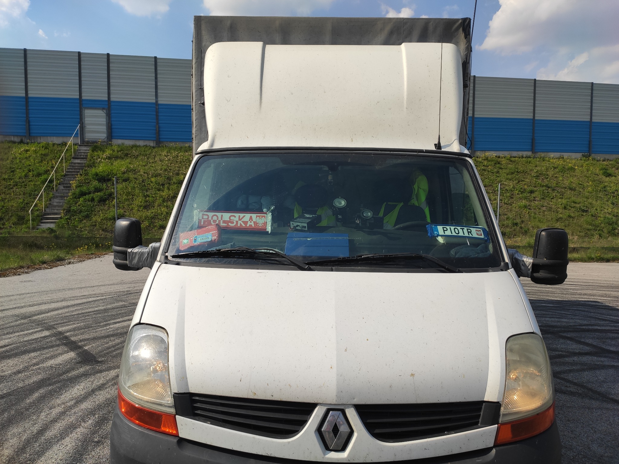 Przód białego busa Renault z tabliczkami 'Polska' i 'Piotr' na desce rozdzielczej, zaparkowanego na asfaltowym placu z widocznymi śladami opon, w tle ekran akustyczny i zielony nasyp.