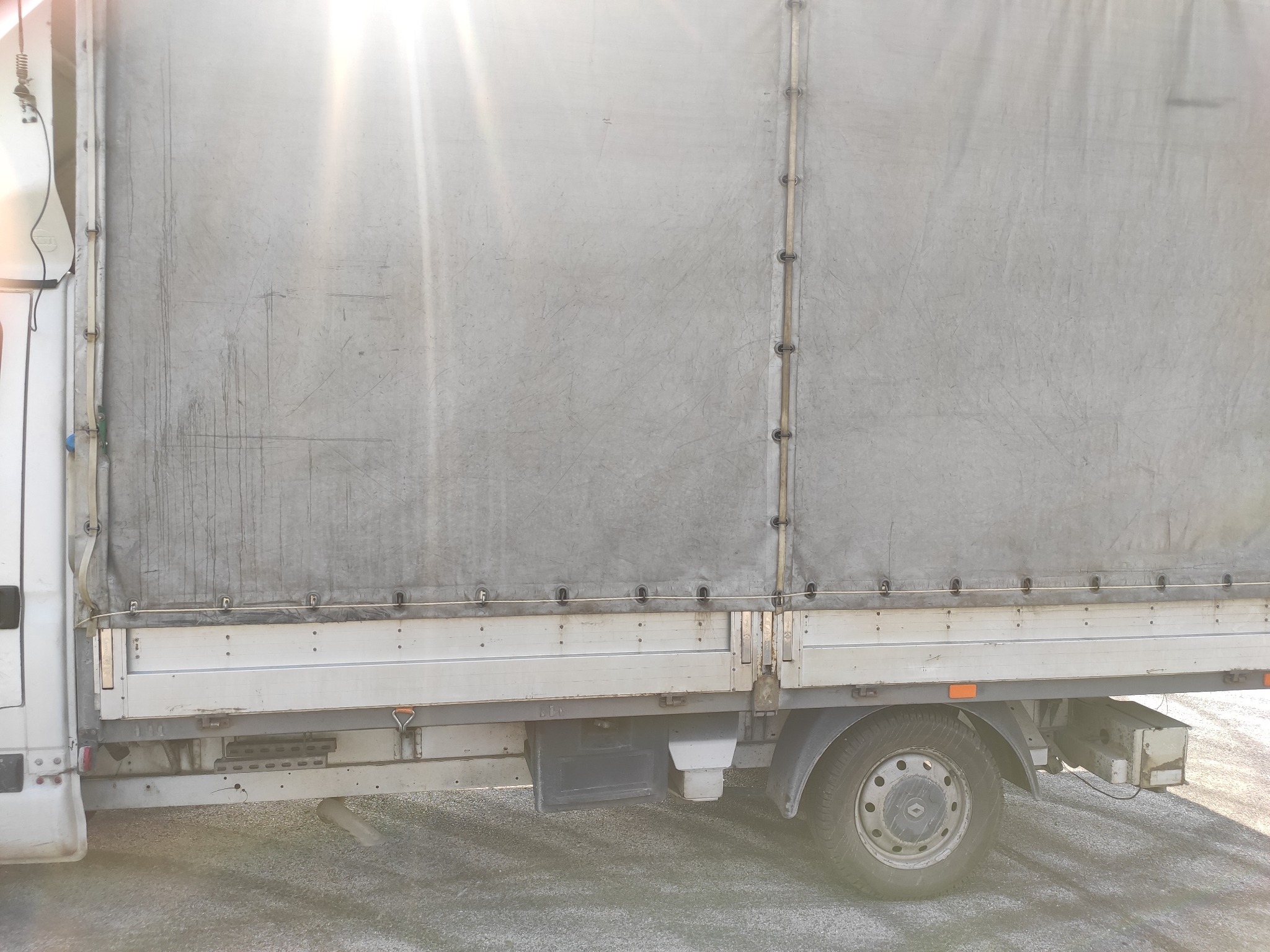 Bok pojazdu dostawczego z plandeką, widoczne mocowania plandeki i koło, oświetlone mocnym słońcem.