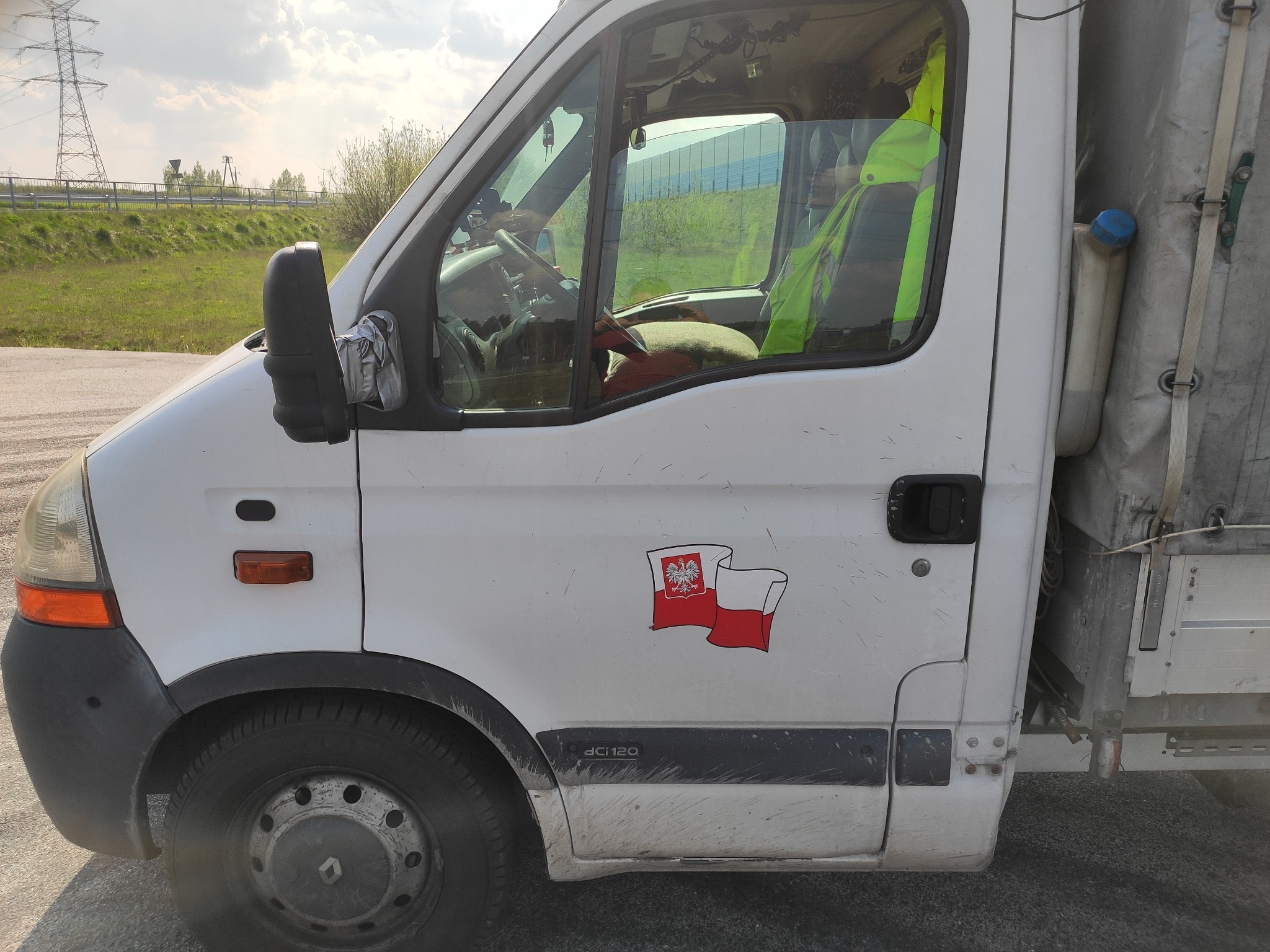 Biały, lekko zabrudzony bus z naklejką flagi Polski na drzwiach, zaparkowany przy drodze z widoczną w tle linią wysokiego napięcia.