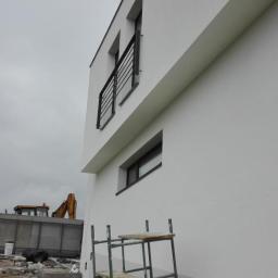 Kontrast - Fragment elewacji budynku z pomalowaną na biało górną częścią i szarą dolną, widoczne rusztowanie, okna z czarnymi ramami, balkony z czarną balustradą oraz koparka w tle.